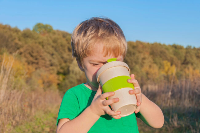 The boy travels near the forest. Sunset light. He drinks from the eco cup